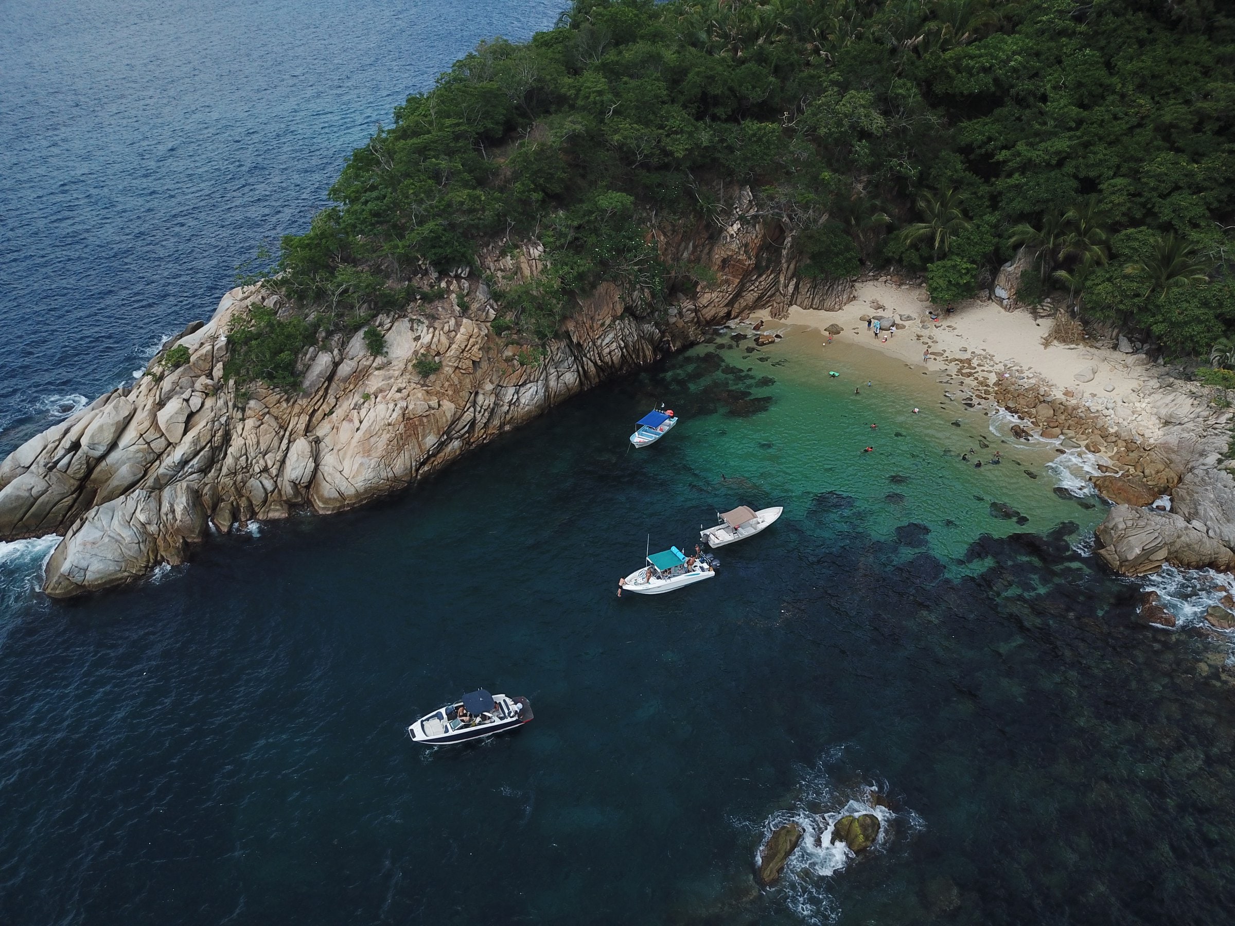 Una cala de la bahía desde el aire: roca, selva, una playa pequeña y tres lanchas ancladas.