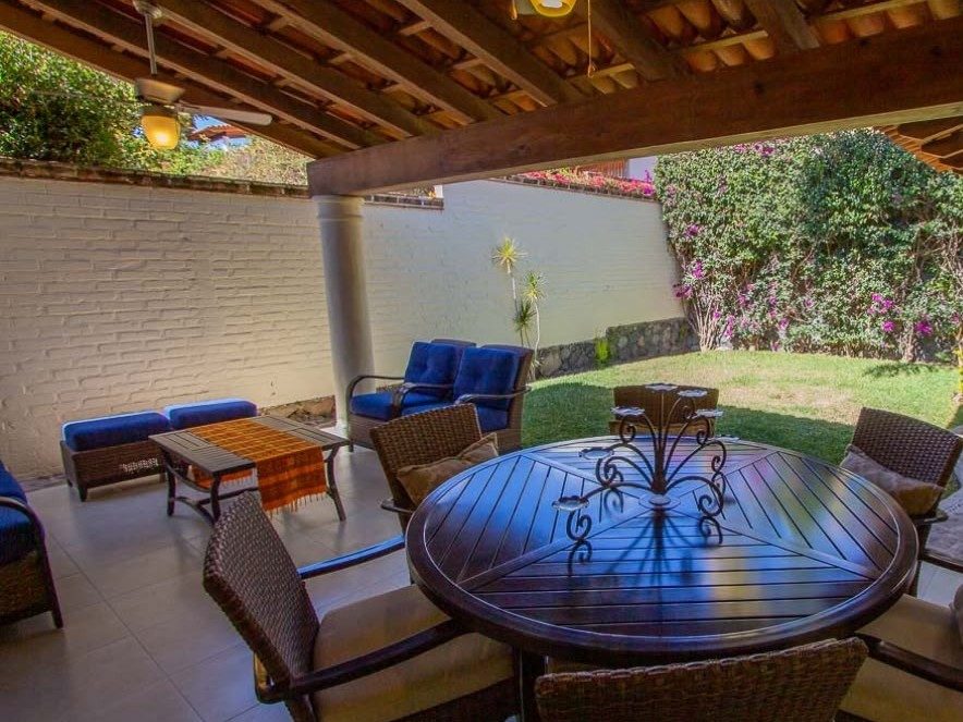 La terraza techada: Mesa redonda y sala bajo vigas de madera, junto al jardín.